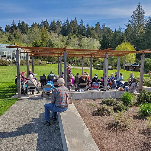 A diverse group of individuals enjoying a sunny day together in Marie’s Terrace park, seated on benches and grass.